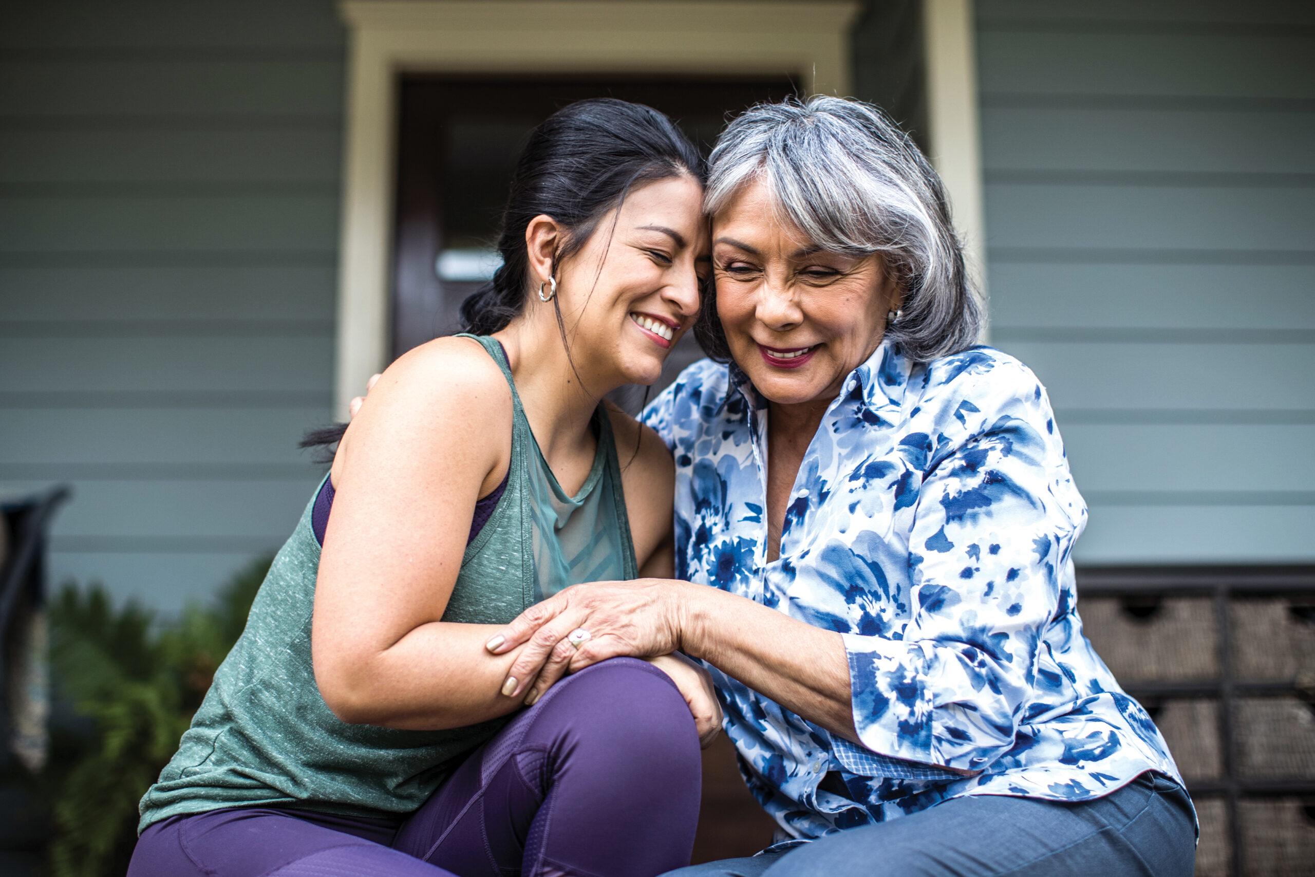 A middle-aged woman embracing and leaning her head gently against an elderly woman sitting on a porch, both smiling in affection.