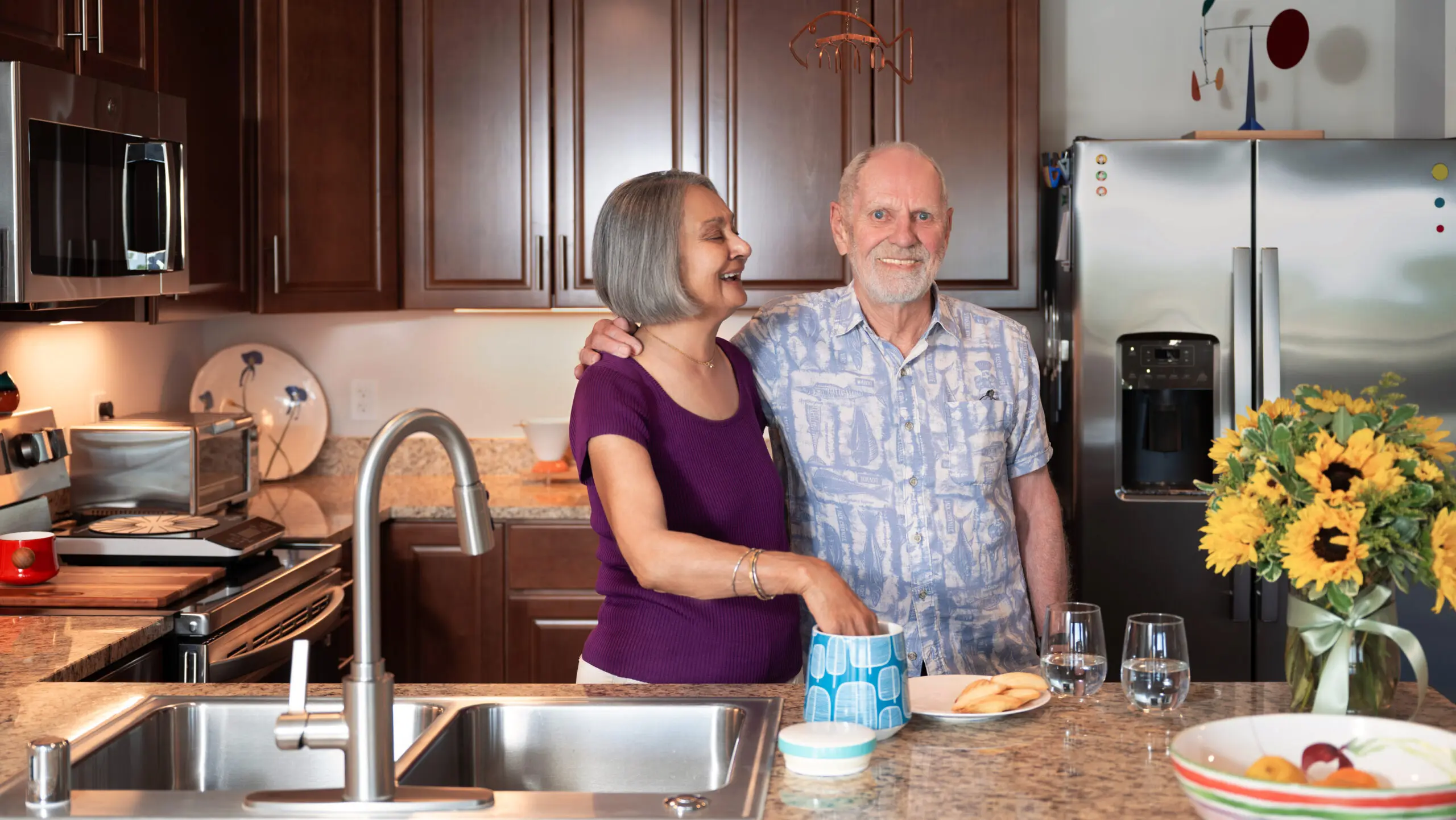 An older couple smiling in a modern kitchen as the woman reaches into a blue-patterned cookie jar on the marble countertop.
