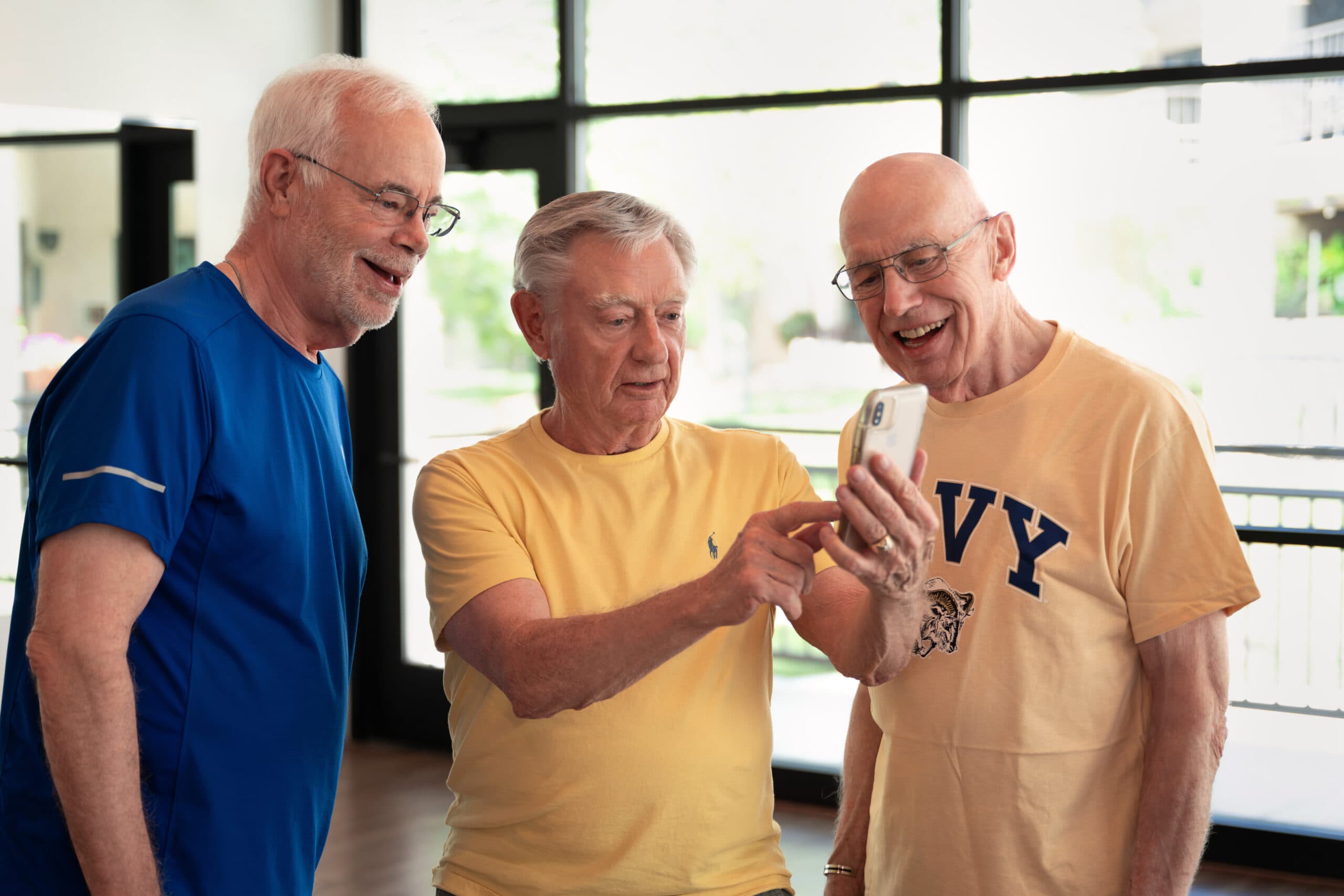 Three older men gathered indoors looking at a smartphone together, one pointing at the screen as they share a laugh.