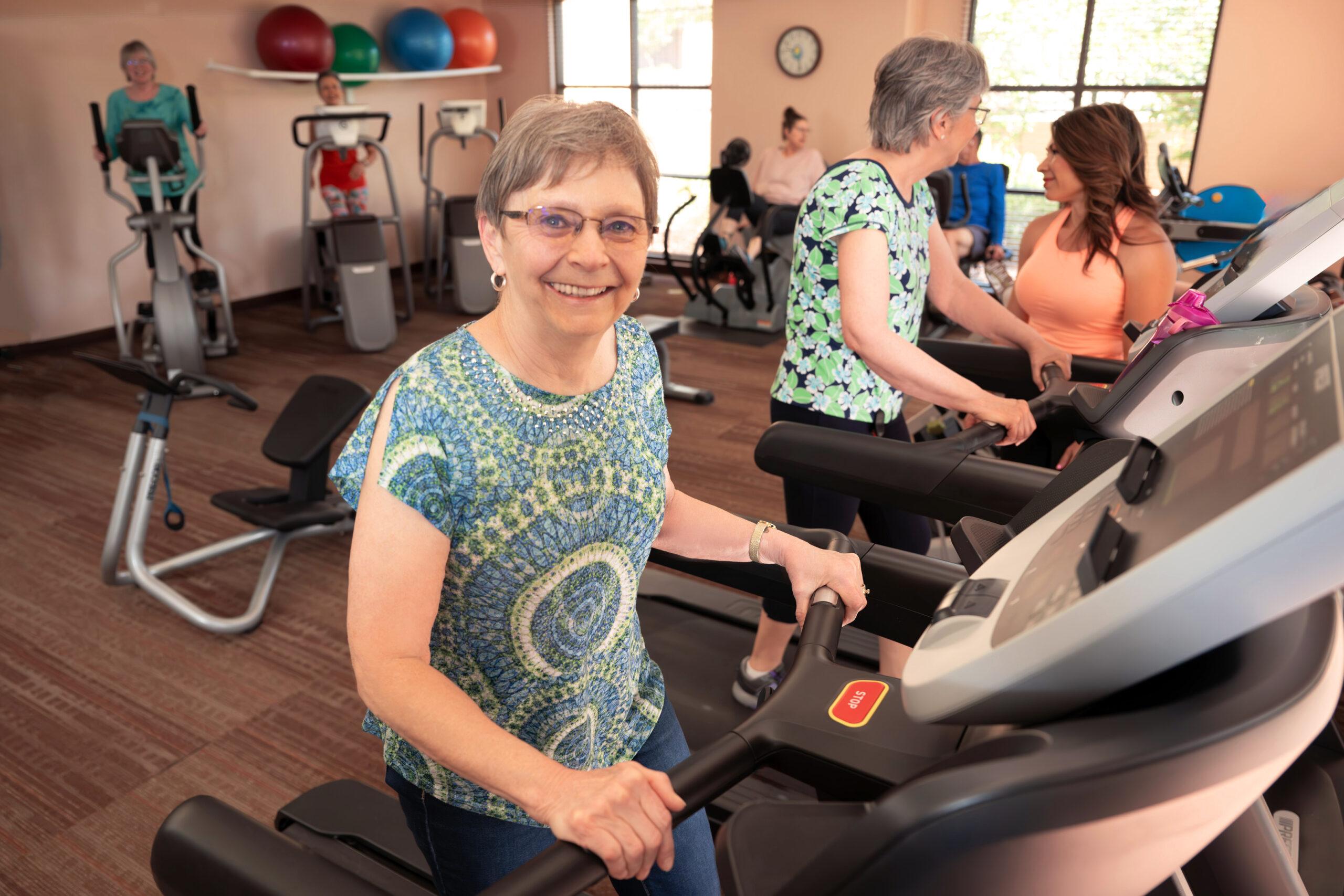 An older woman walking on a treadmill in a gym, smiling as other members exercise in the background.