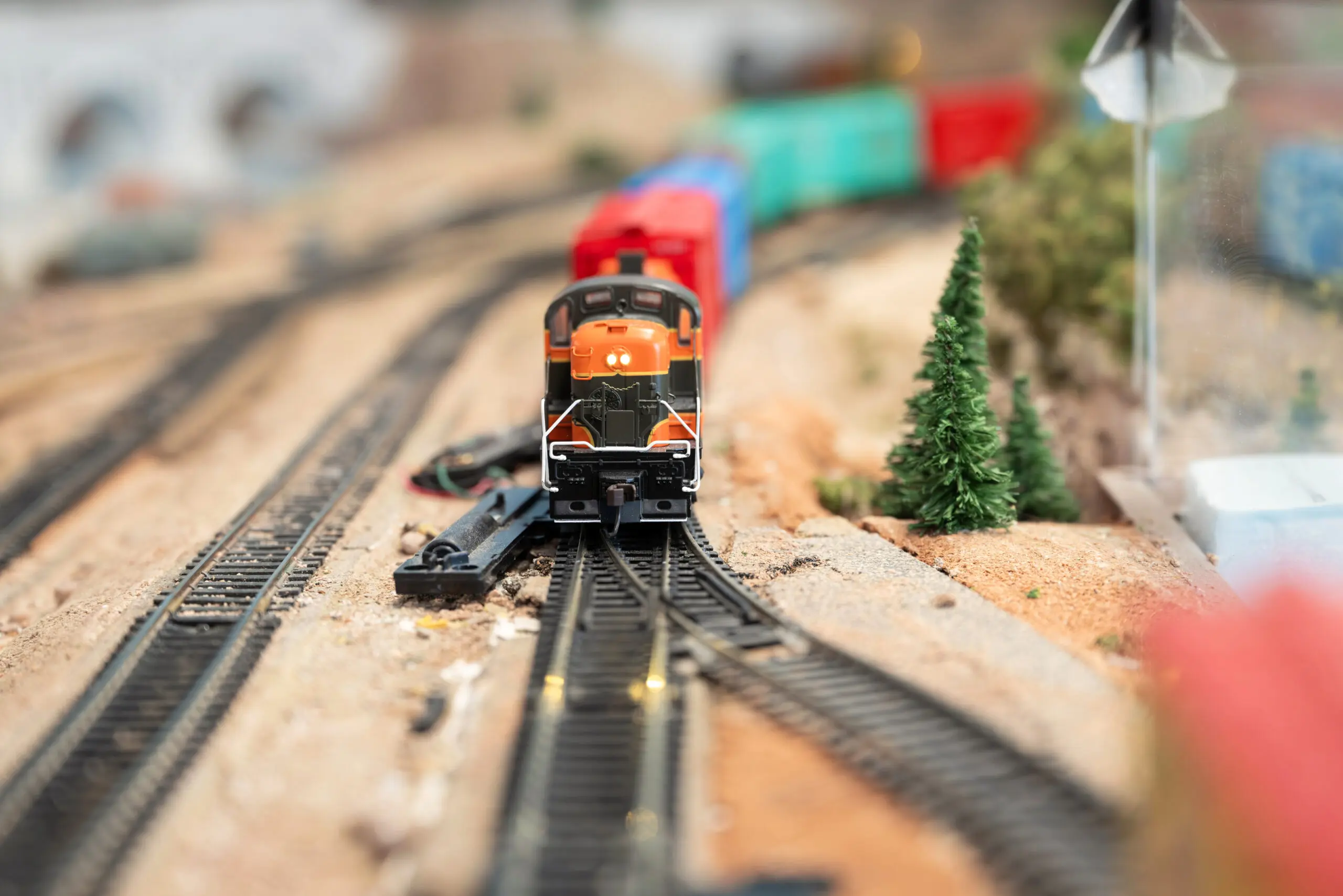 Close-up of an orange model train locomotive on miniature tracks with blurred freight cars and scenery behind.