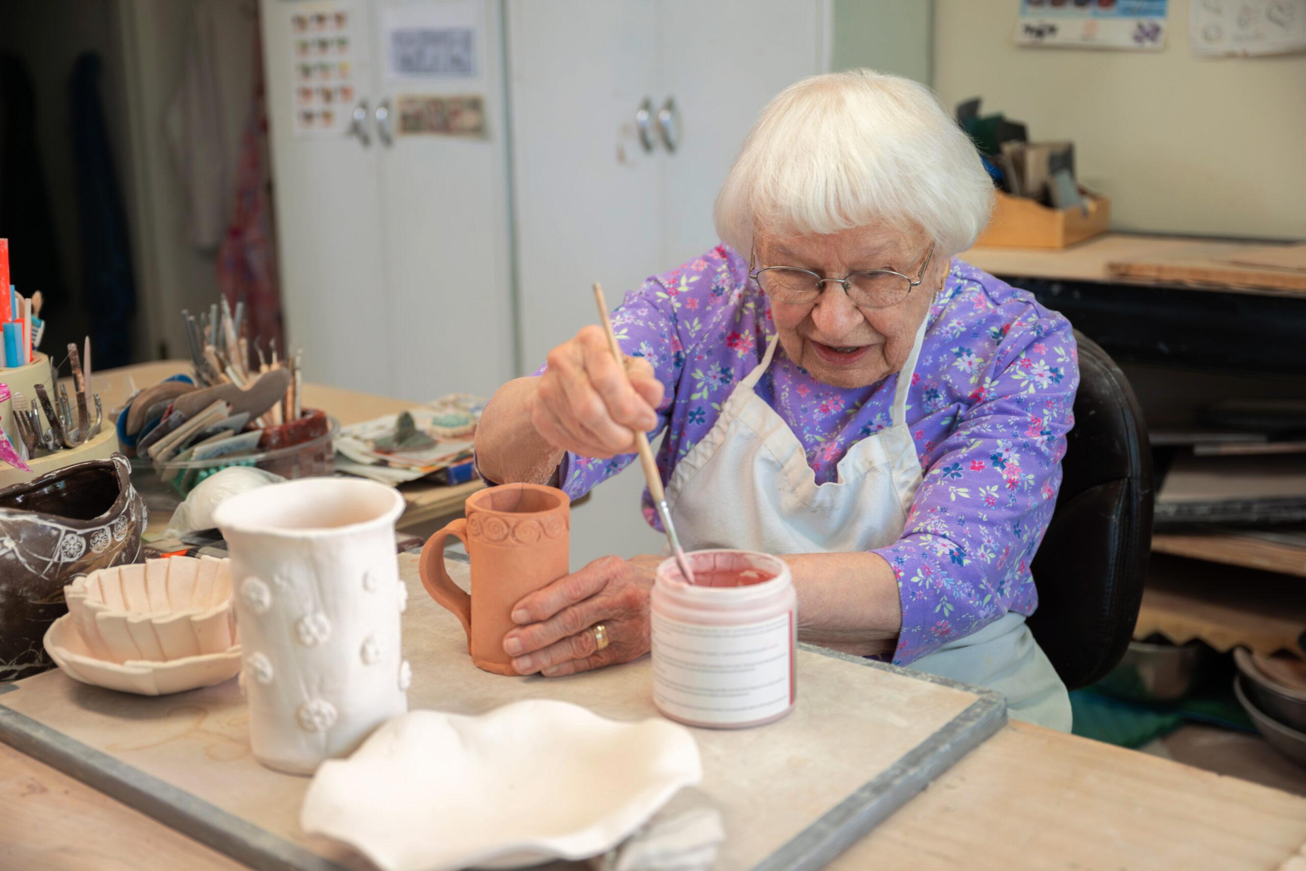 An elderly woman in a floral blouse painting a ceramic mug with pink glaze at a pottery studio workstation.