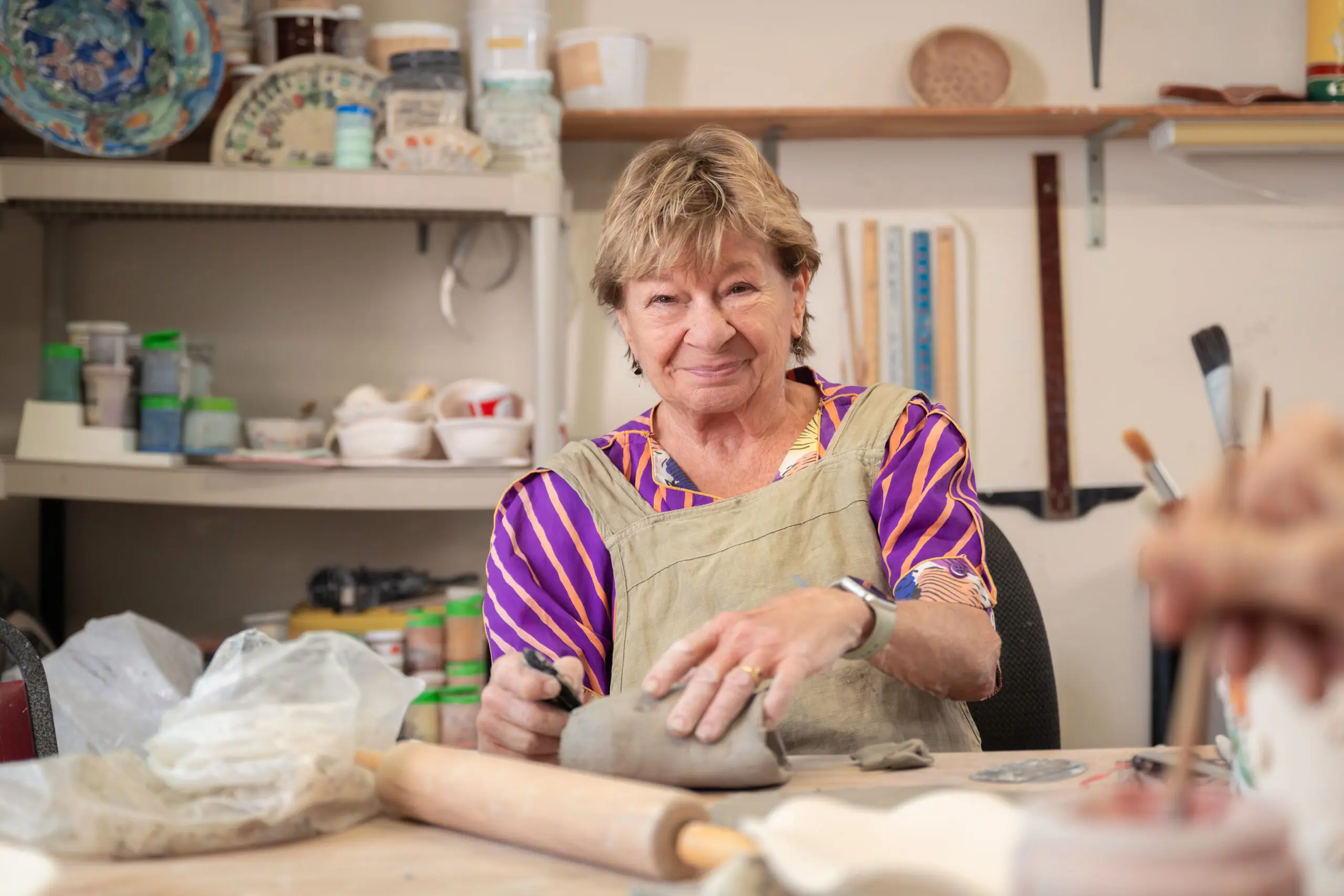 An elderly woman sitting at a pottery studio table, shaping a lump of clay with a tool, surrounded by shelves of glazes and ceramics.