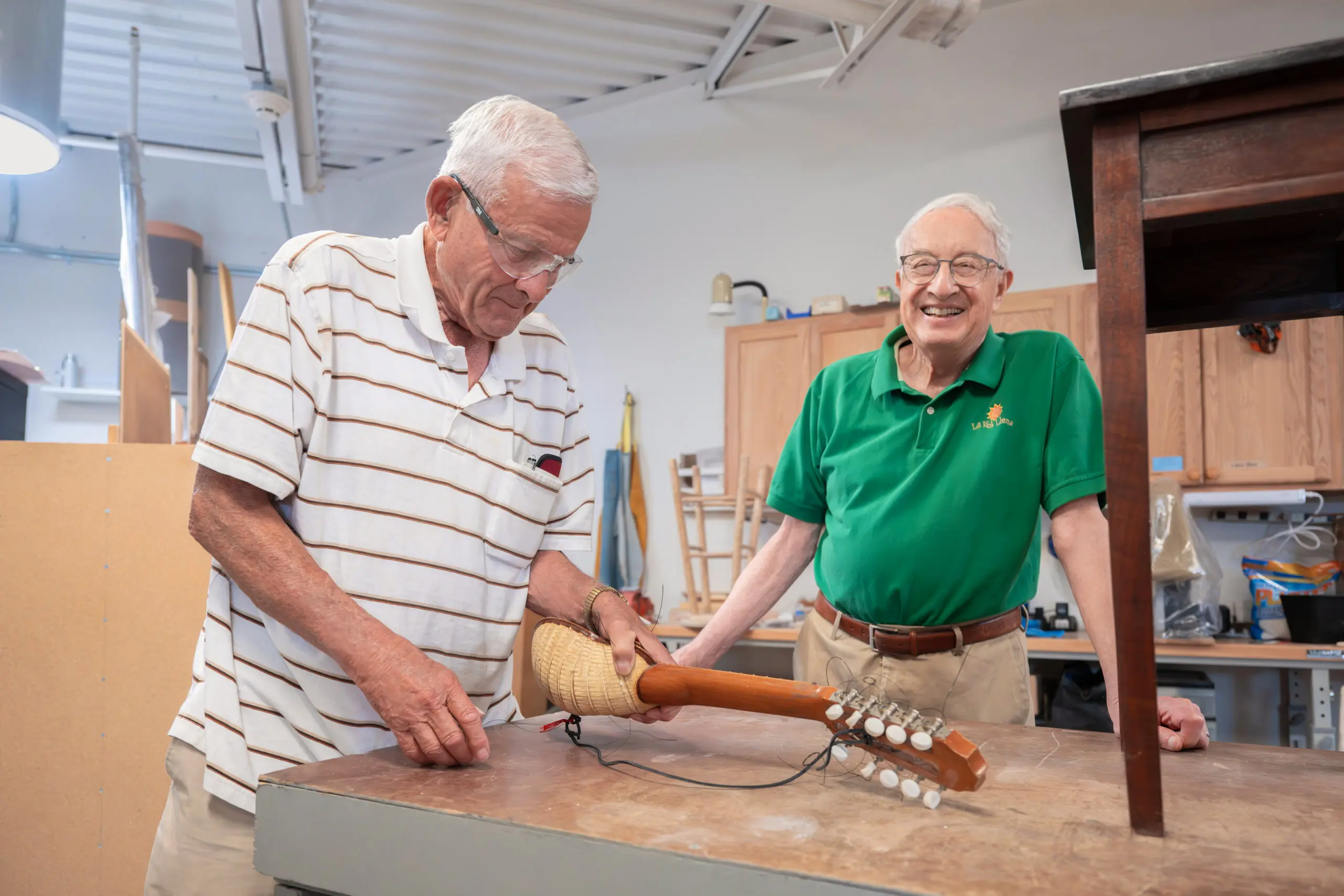 Two senior men at a workshop bench, one tuning the strings of a mandolin-like instrument while the other watches and smiles.