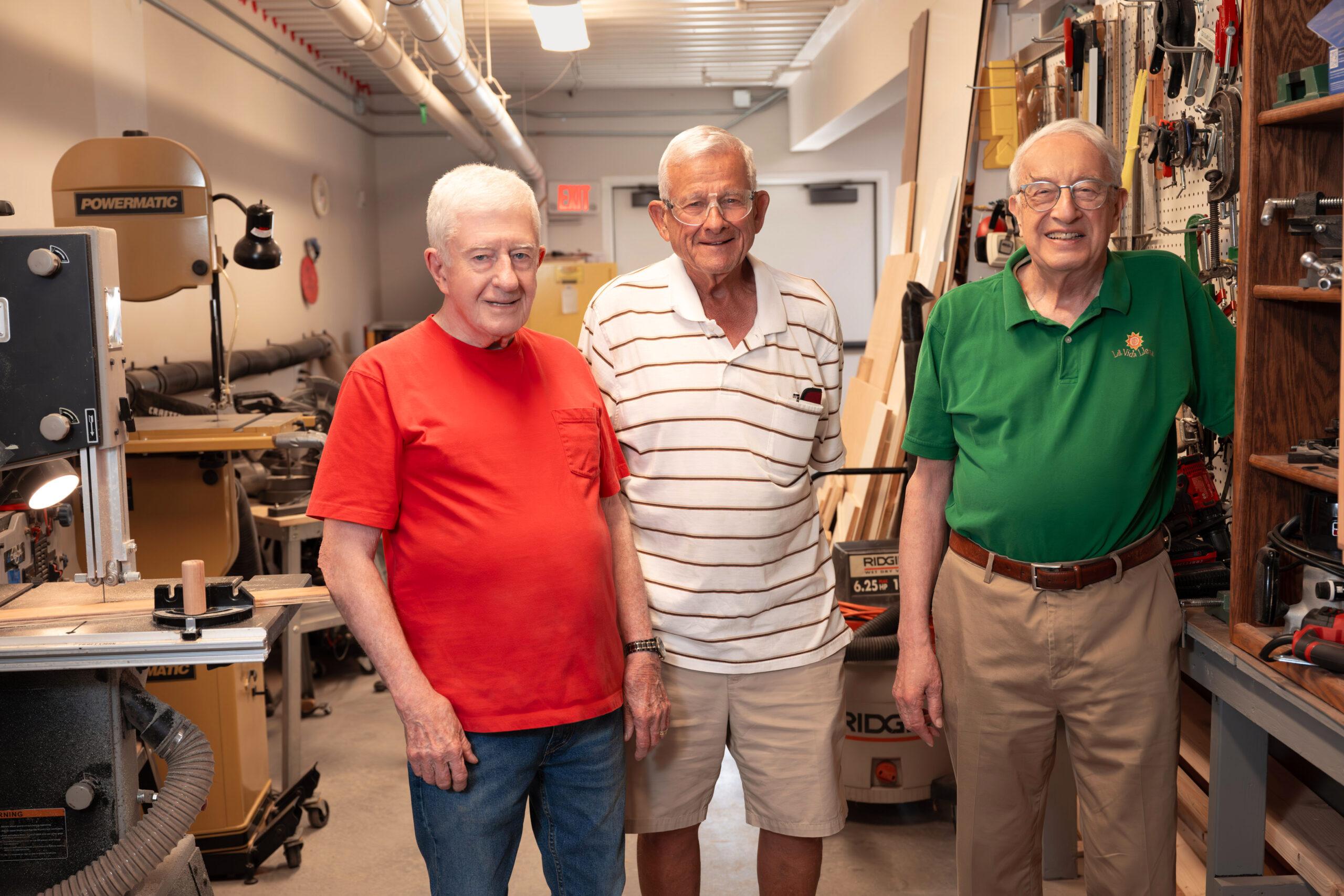 Three elderly men standing side by side in a woodshop, posing together with woodworking machinery and tools visible around them.