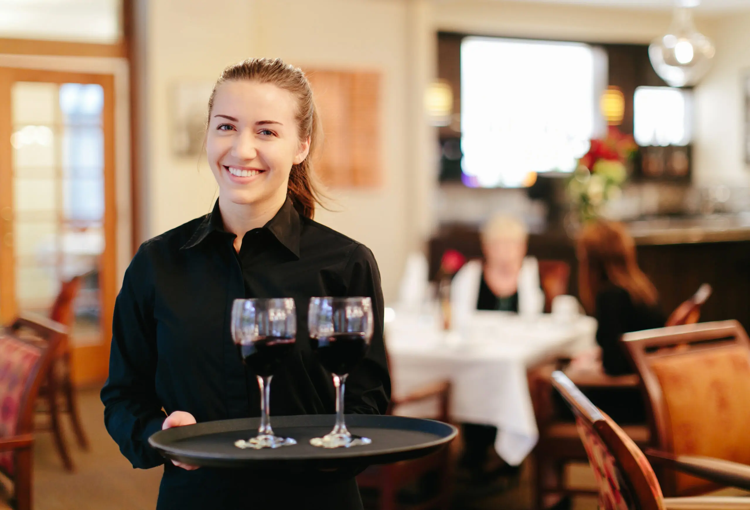 A smiling server in a black shirt carrying a round tray with three glasses of red wine in a warmly lit dining room.